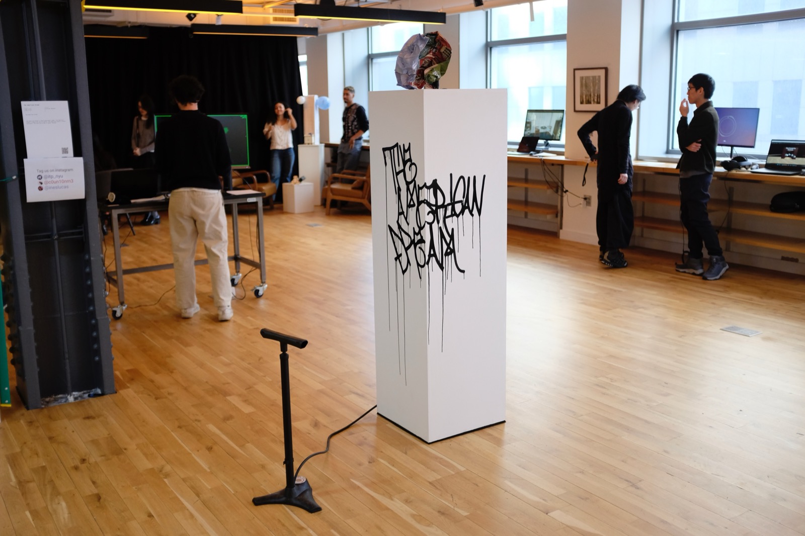 The full installation at the ITP winter show: a white pedestal tagged with dripping black graffiti reading The American Dream, a crumpled balloon on top, and a bicycle pump standing in front