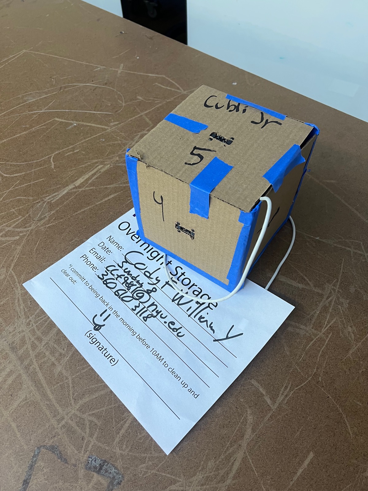 Cardboard cube prototype labeled Cubli Jr, taped with blue tape, on an overnight-storage form