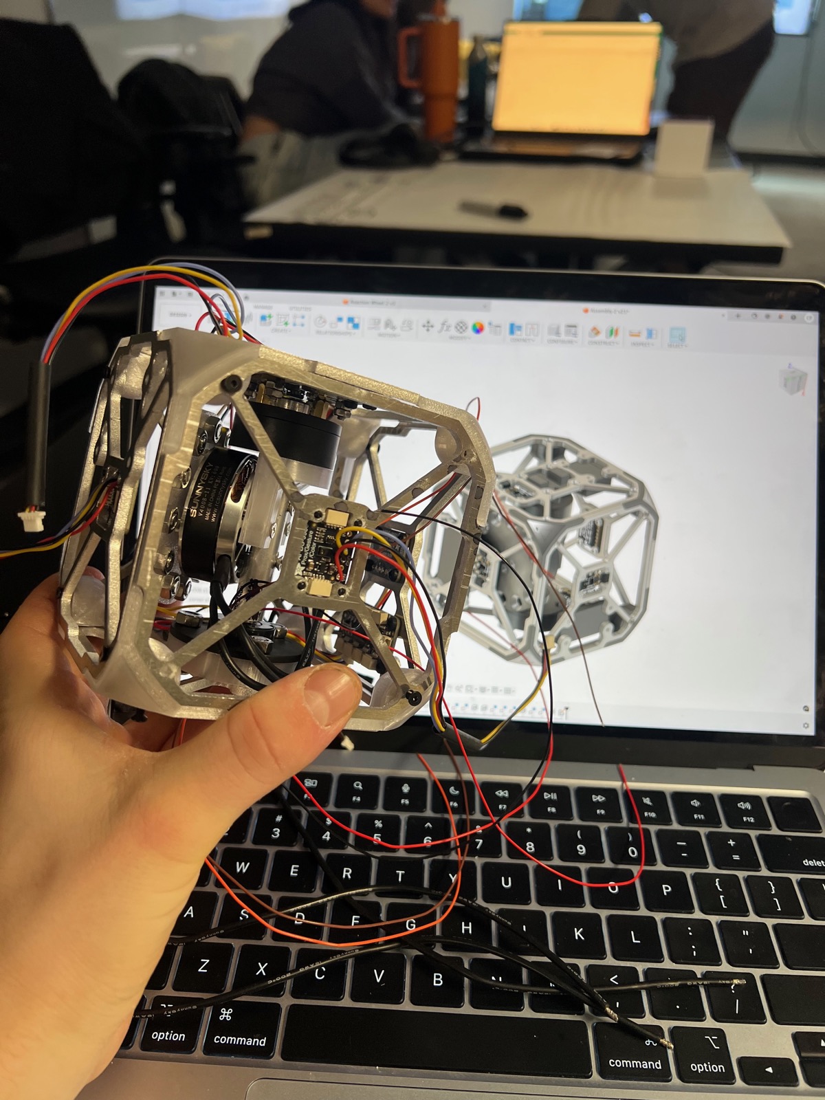 Hand holding the aluminum-frame cube prototype in front of a laptop showing its CAD model