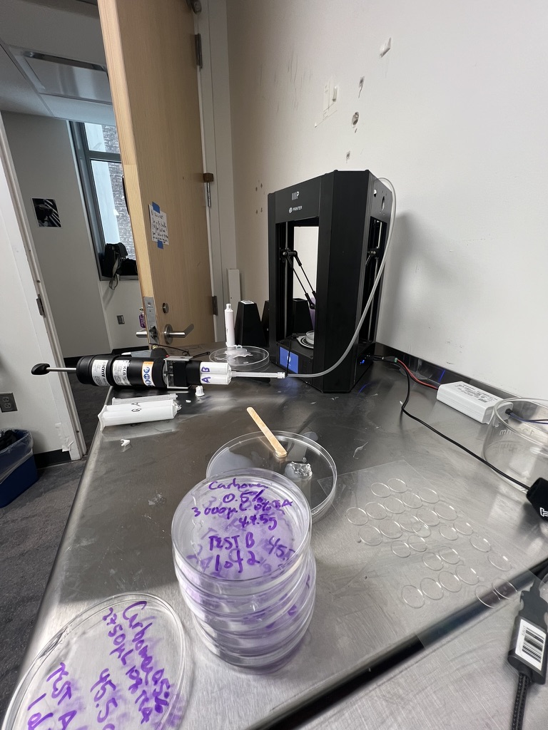 A steel work table with a delta 3D printer, a pressure dispensing gun feeding it by tube, and stacks of labeled gel test petri dishes