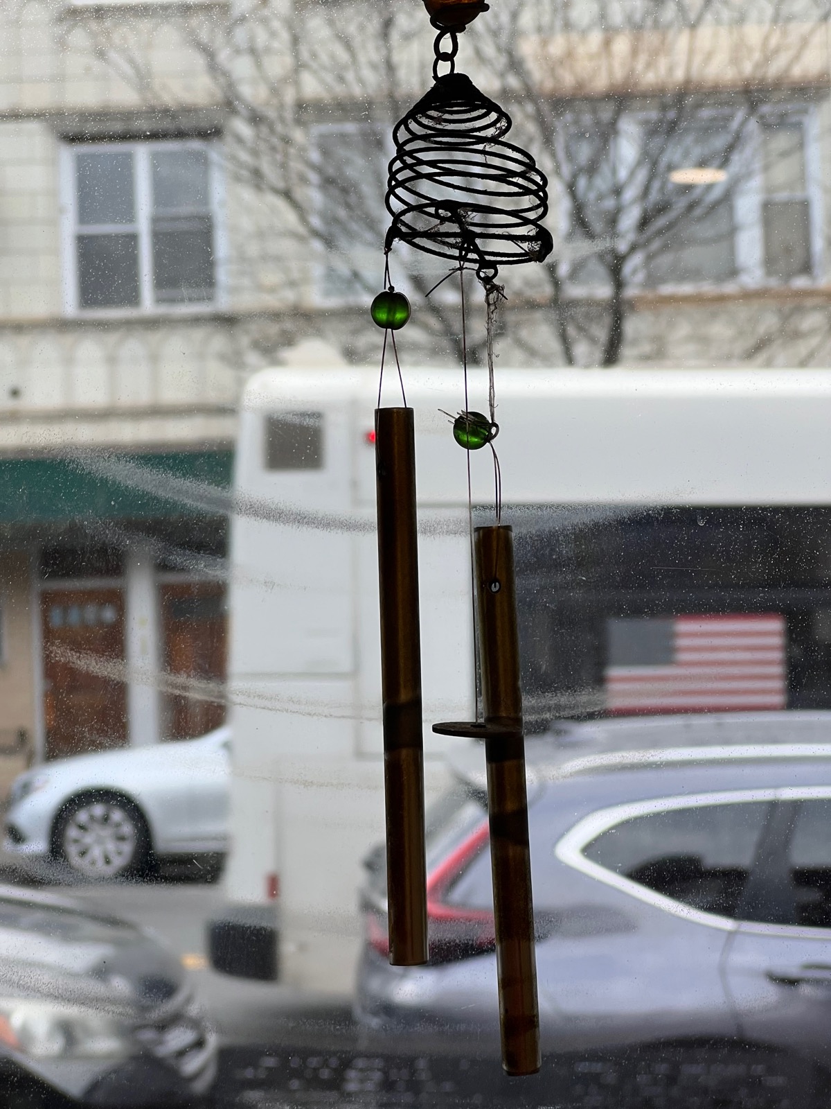 Two aged brass wind chime tubes hanging from a black wire spiral, with green glass beads, in front of a window looking onto a blurry street with a bus and parked cars.