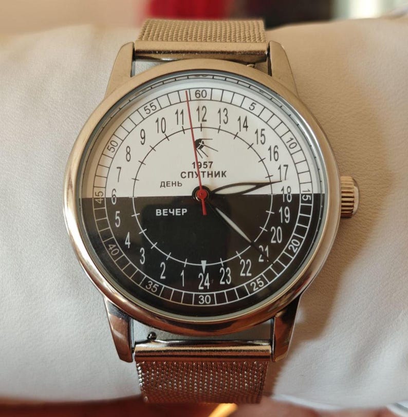 May include: A silver wristwatch with a unique black and white face. The watch face is divided into day and night sections, with the text "1957 Sputnik" and Cyrillic text. The watch has a mesh band.