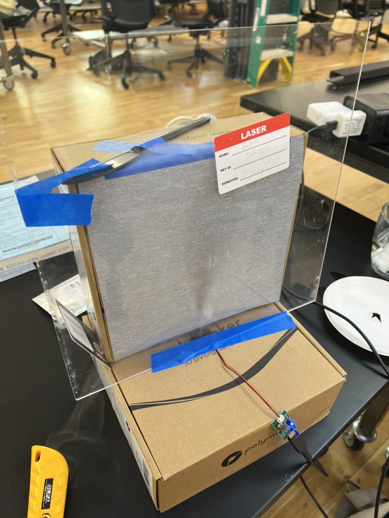 Test rig on a shop table: a sheet of gray calligraphy paper taped to a cardboard box, a clear acrylic sheet in front of it, and a small ultrasonic mister board glowing blue on the box below.