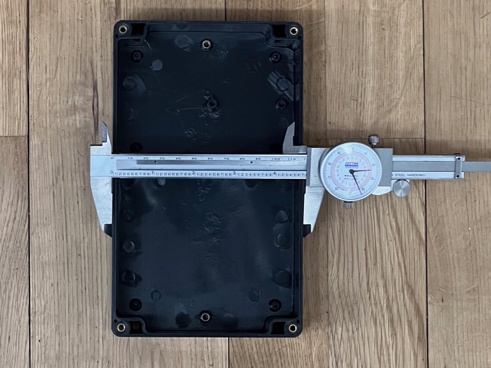 The empty black waterproof enclosure on a wood floor with a dial caliper clamped across its width.
