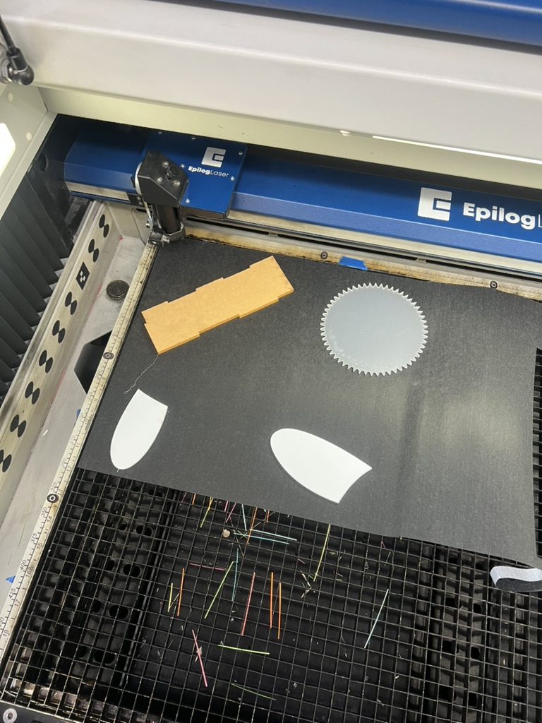 Dark calligraphy paper on the bed of an Epilog laser cutter, held flat by white acrylic scraps and an MDF strip, with cut offs scattered on the grid below.