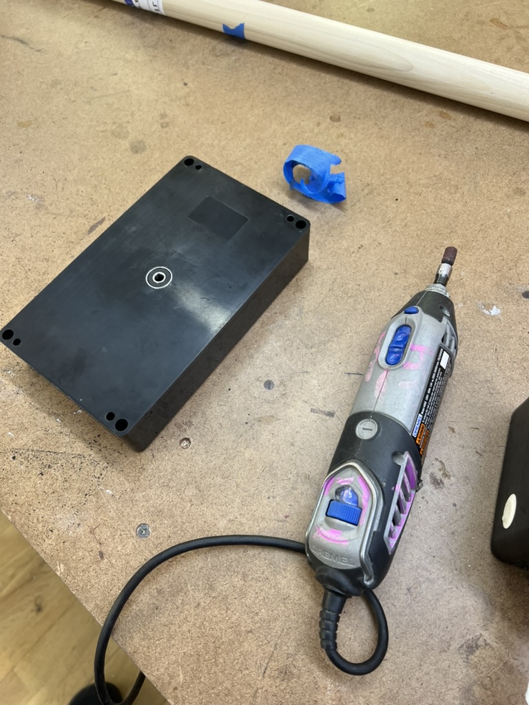 The black enclosure on a workbench with a freshly drilled hole in its center, next to a Dremel with a sanding drum and a curl of blue tape.