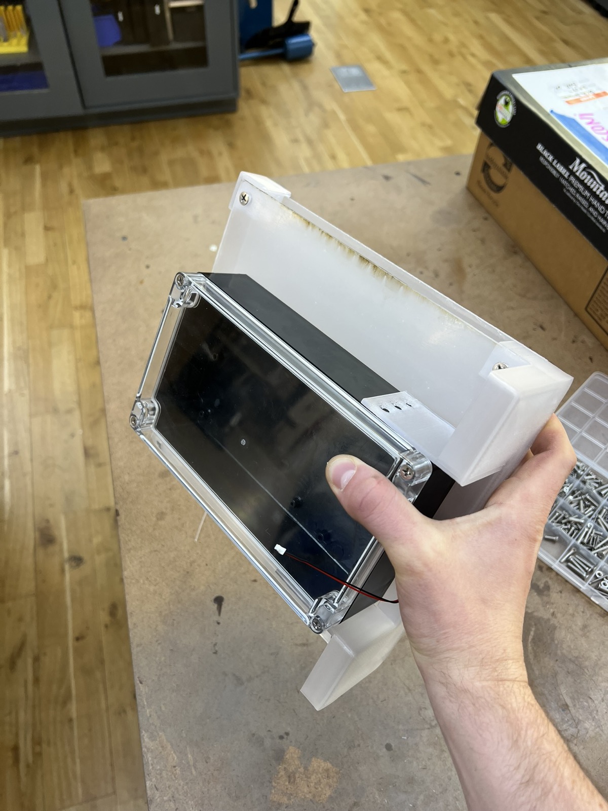 A hand holding the black enclosure with its clear lid, mounted on a frosted acrylic panel with printed brackets at each end and a laser browned edge along the top.