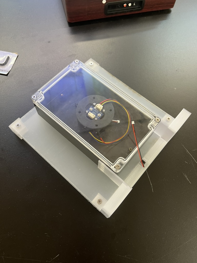The enclosure with its clear lid screwed onto the frosted acrylic panel on a black table, the printed encoder mount and wiring visible through the lid.