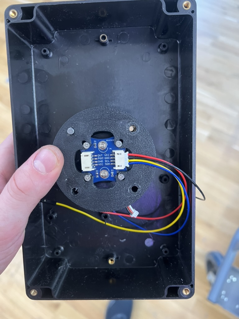 The inside of the empty black waterproof enclosure, with a round 3D printed mount at its center holding a small blue magnetic encoder board wired in red, yellow, blue and black.