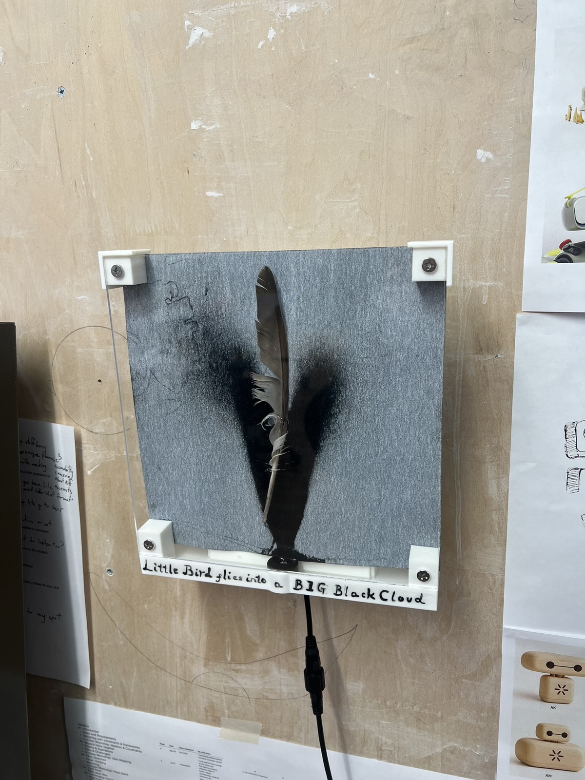 The finished piece hanging on a plywood wall: gray calligraphy paper behind clear acrylic in white printed brackets, a dark feather standing at the center inside a V of black mist, and Little Bird flies into a BIG Black Cloud hand lettered along the base, with a power cord hanging below.