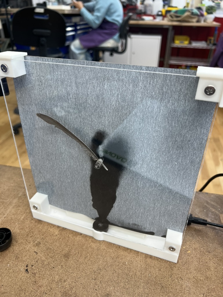 The first full assembly on a workbench: gray calligraphy paper behind clear acrylic in white printed brackets, a dark feather on the acrylic rod at its center like a clock hand, and a black mist stain rising from the mister below.