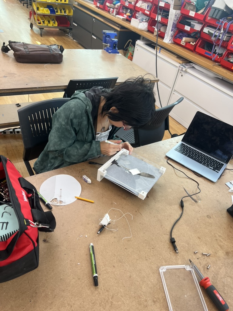 Kezia bent over a shop table, hand lettering the white base of the piece with a paint pen, with a laptop and tools on the table.