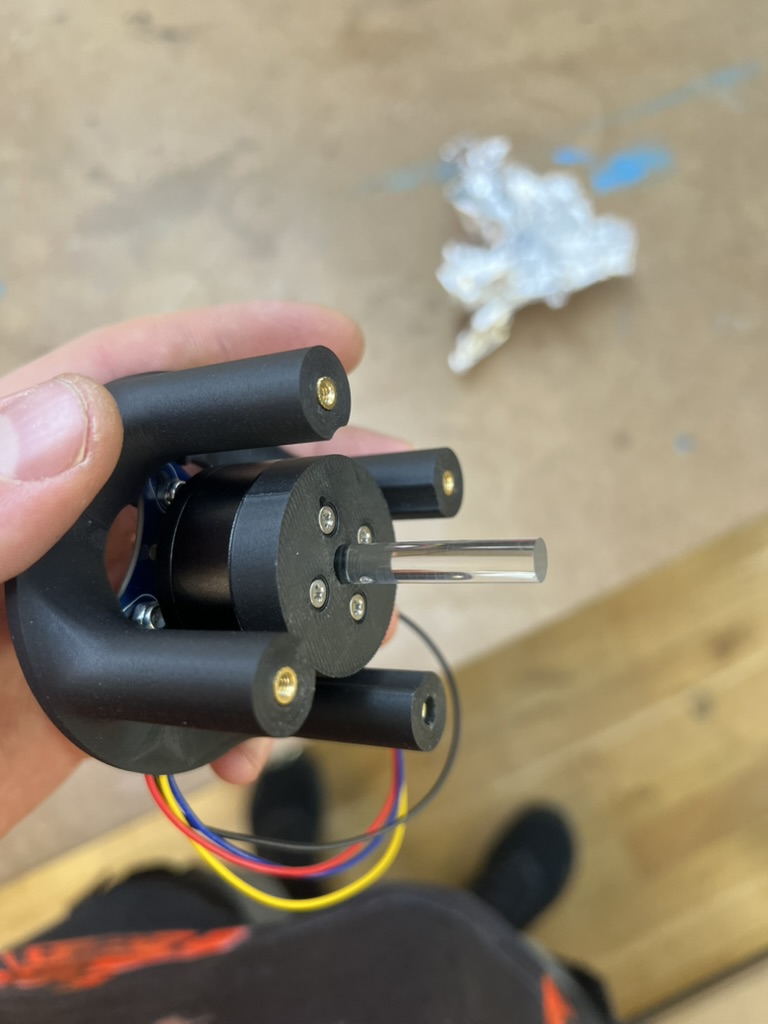A hand holding the black 3D printed motor mount, four posts with brass heat set inserts around the motor, a short clear acrylic rod sticking out of its center and colored wires hanging below.