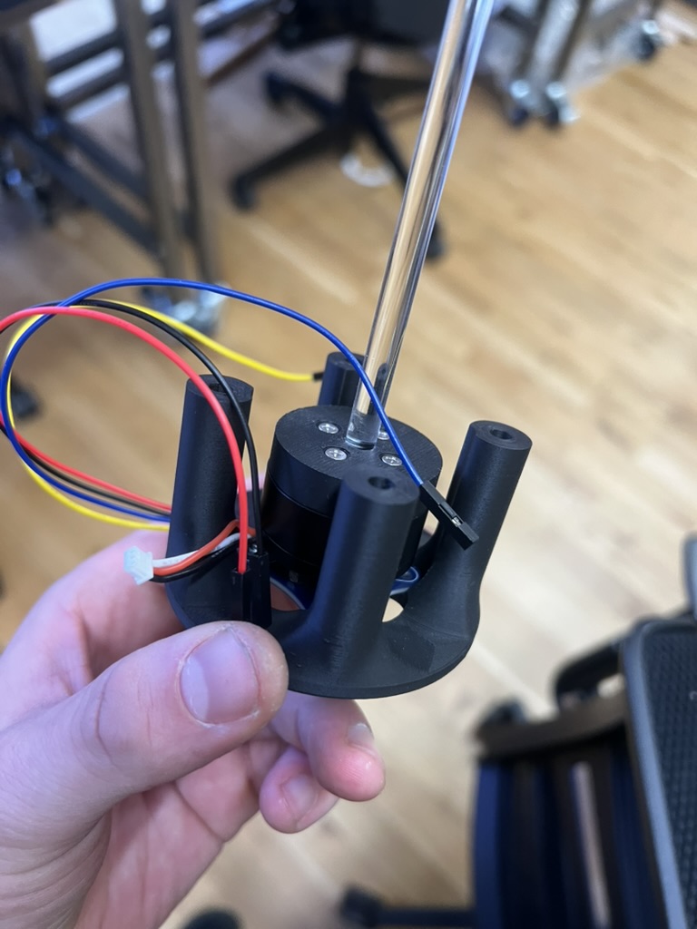 The motor seated in its printed mount, held in hand, with a long clear acrylic rod rising from the rod grab and jumper wires looping out the side.