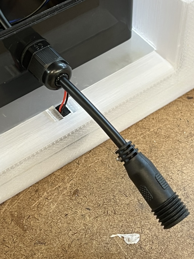 A waterproof cable gland on the side of the enclosure with a short black 12v lead ending in a screw on connector, its red and black wires running into the white base tray.